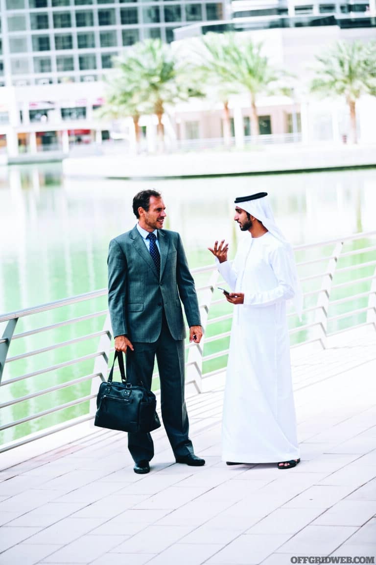 Two adult males from different cultures practice building rapport by having a conversation next to a watery area.
