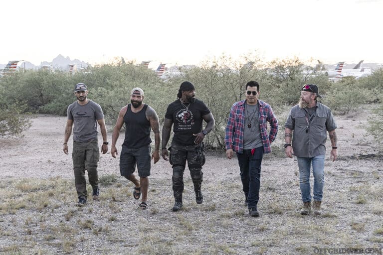 Photo of Protector Symposium cadre walking side-by-side through an opening in scrub brush with an airfield in the background.
