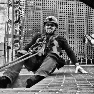 Black and white photo ofJoey Nickischer sitting on cobble stones and rigged up with technical rope gear.