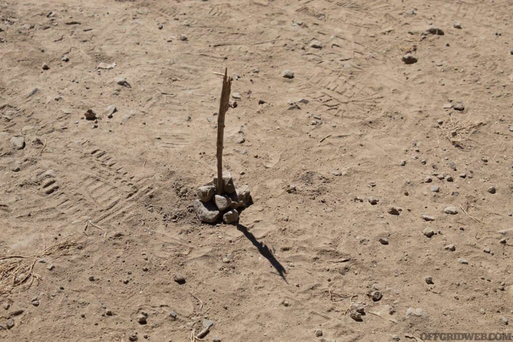 Photo of a single straight stick stuck into the dusty ground in full sunlight. The stick is casting a shadow and will be used as the starting point for an improvised sundial. Its a useful way of navigating without tools.