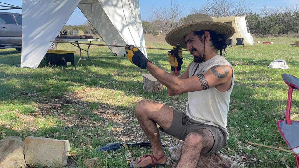 Photo of a primitive wilderness survival student eyeing the length of an arrow to check for straigness.