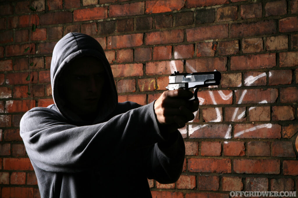 Faceless adult in a hoody in a dark alley pointing a gun at something outside of the frame.