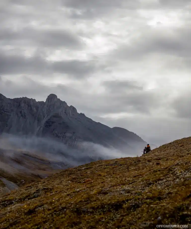 Photo of an adult male sitting on a foothill, lookin at snow covered and cloud shrouded mountains in the background.