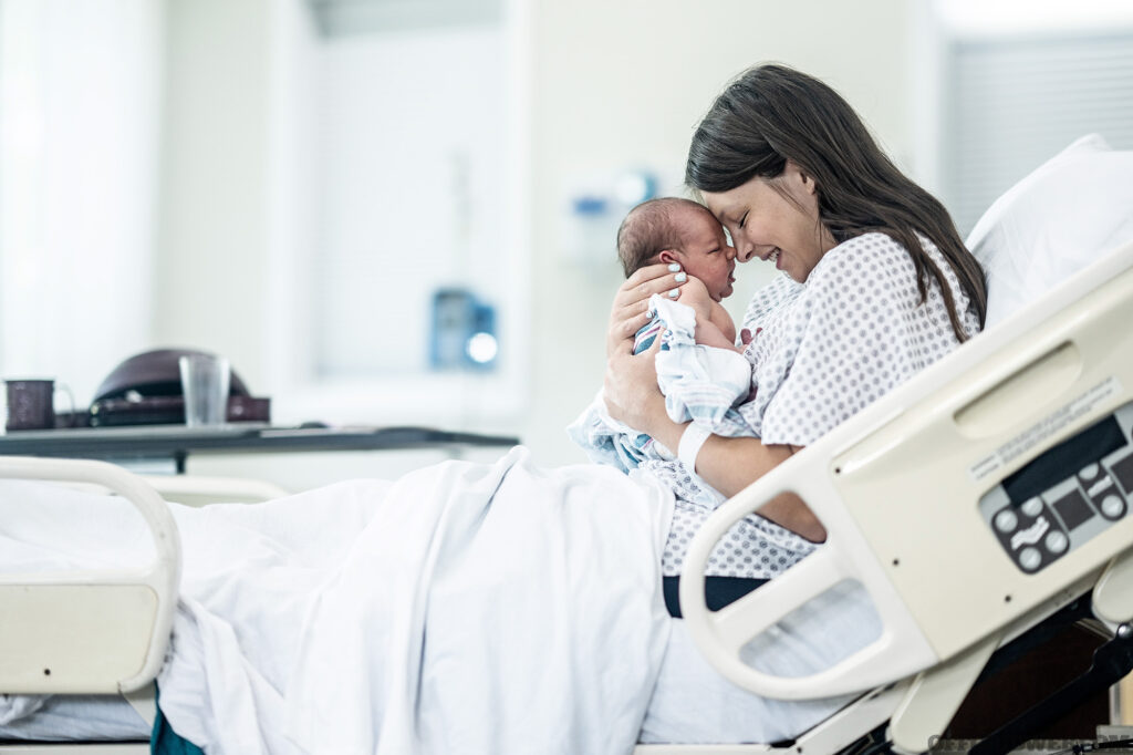 A new Mother sits up in her hospital bed shortly after delivery as she holds her newborn out in front of her and studies his features. She is wearing a hospital gown and is laying in her hospital bed with the infant.