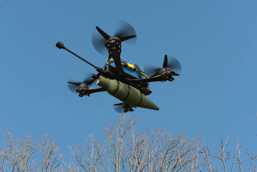 A combat drone flies over the trees, carrying suspended ammunition on a sling